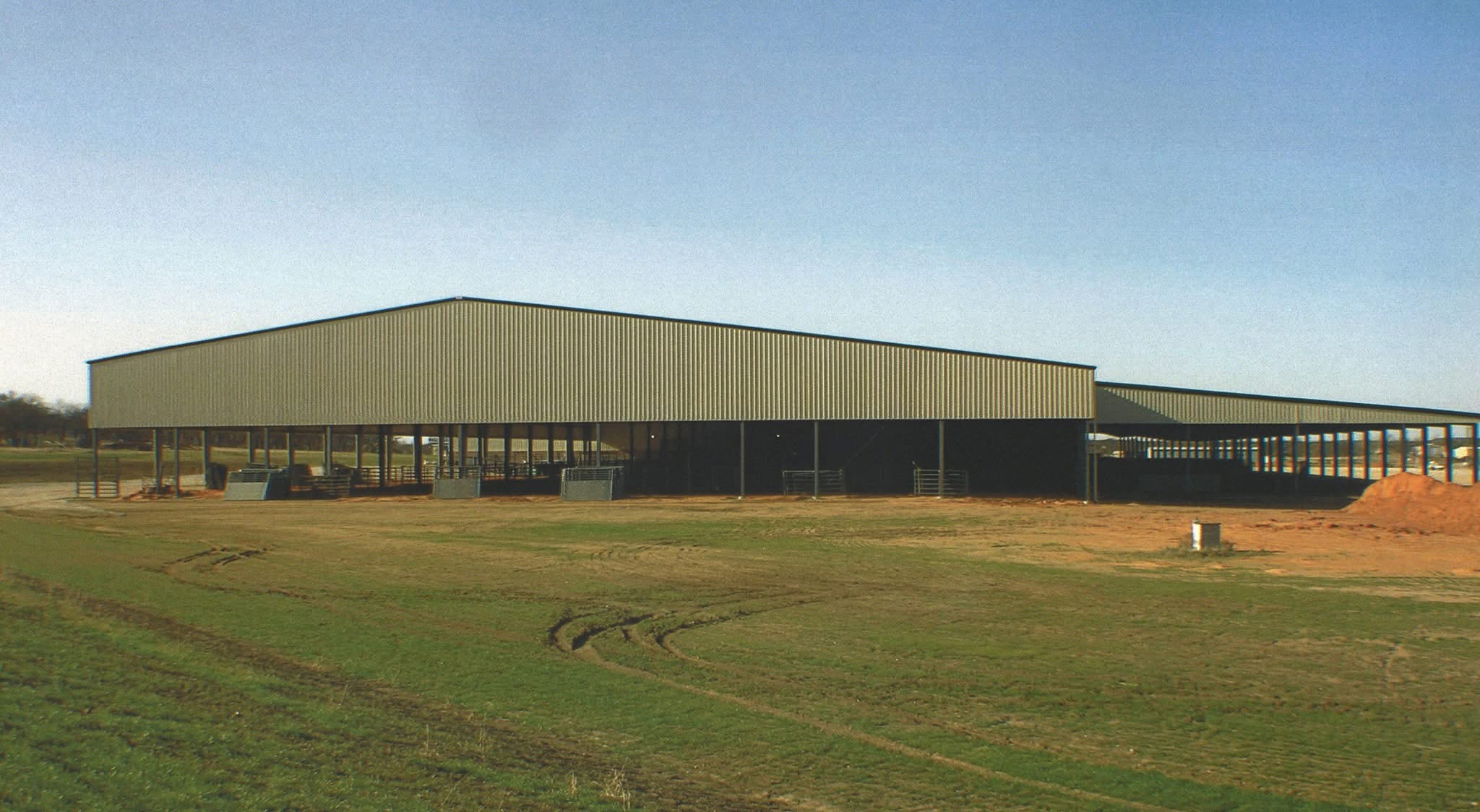 Large open-sided tan steel building on a Texas cattle ranch with pipe fencing