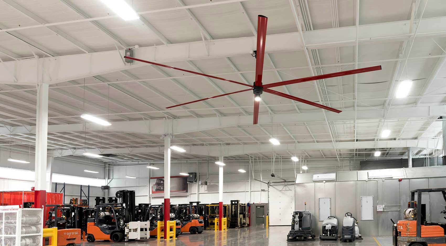 Interior of a steel warehouse with a large red HVLS ceiling fan and forklifts on the floor